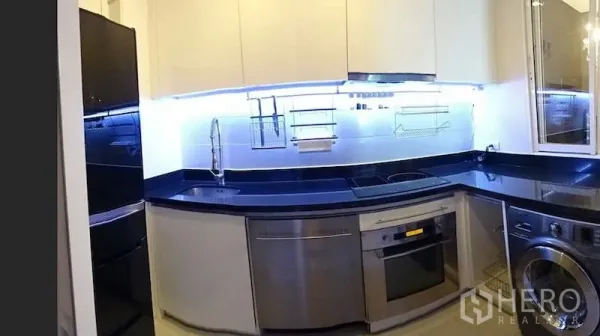 Condominium for rent or sale in Watthana, Bangkok - U-shaped kitchen featuring black stone countertops, stainless appliances and LED backsplash lighting.