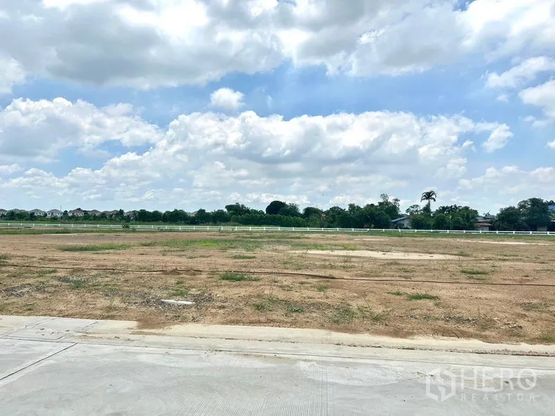 Land for sale in Lam Luk Ka, Pathum Thani - Wide, level land parcel with a white fence and greenery in the distance.