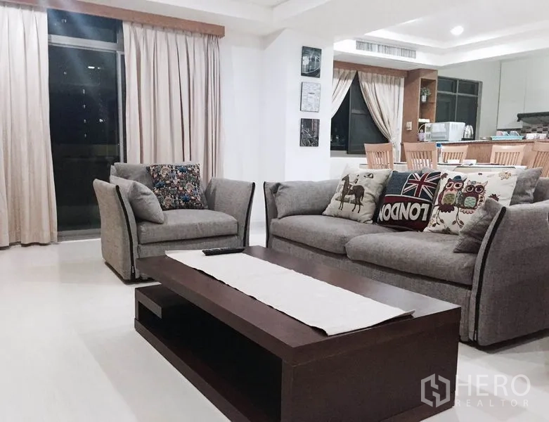 Condominium for rent or sale in Watthana, Bangkok - Living room with sofa set and wooden coffee table beside large curtained windows and a breakfast bar.