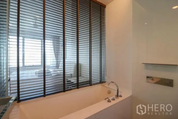 Condominium for rent in Pathum Wan, Bangkok - Modern bathroom with a long soaking tub and window blinds looking into the bedroom.
