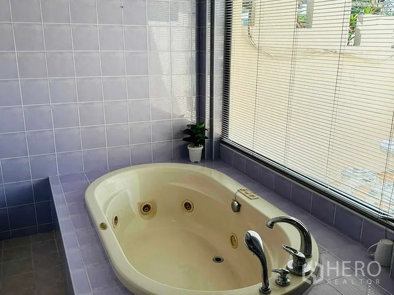 Home Office for rent - Bathroom with oval jacuzzi bathtub beside a window with blinds and lilac tiles.