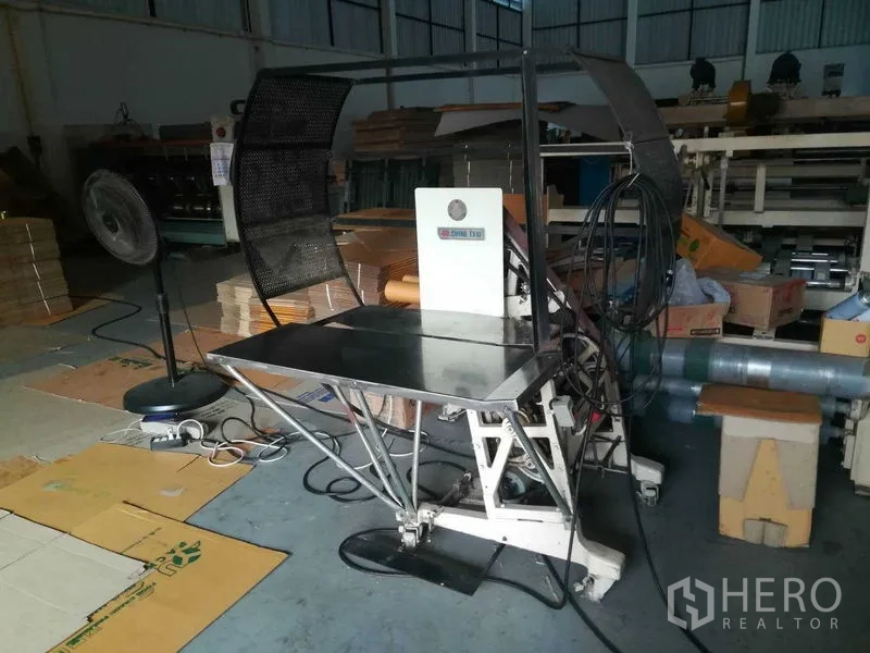 Factory for sale in Bang Len, Nakhon Pathom - Workstation area with equipment, fan and stacked cardboard sheets.