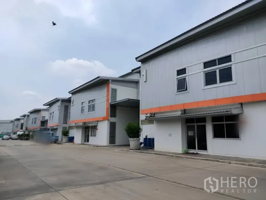 工厂 出租在Phra Nakhon Si Ayutthaya - 工业园内一排现代化厂房，橙色线条点缀，车道宽阔便于通行。