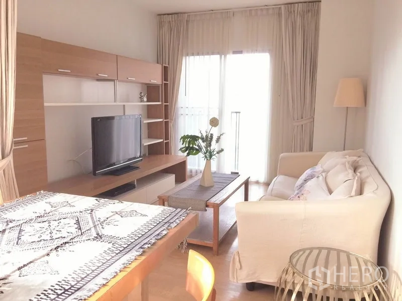 公寓 出租在Bangkok - 明亮客厅，白色沙发、木质电视柜、电视与轻纱窗帘，连通阳台。