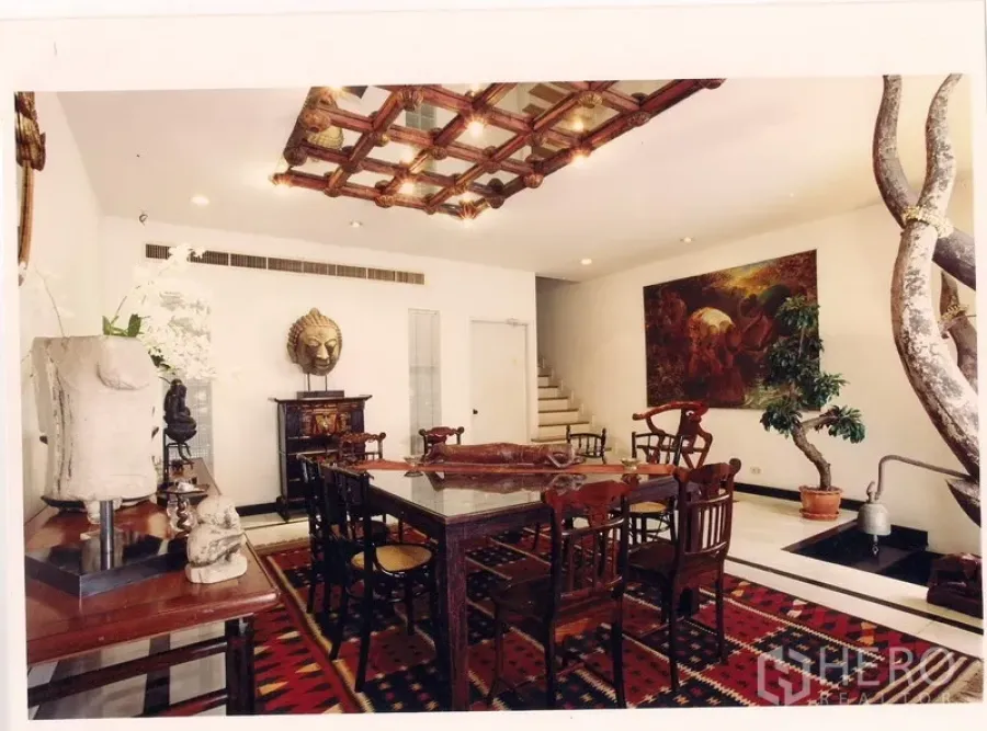 Townhouse for rent or sale in Watthana, Bangkok - Elegant dining room with carved wood ceiling feature, art-lined walls, and staircase beside an indoor water feature.