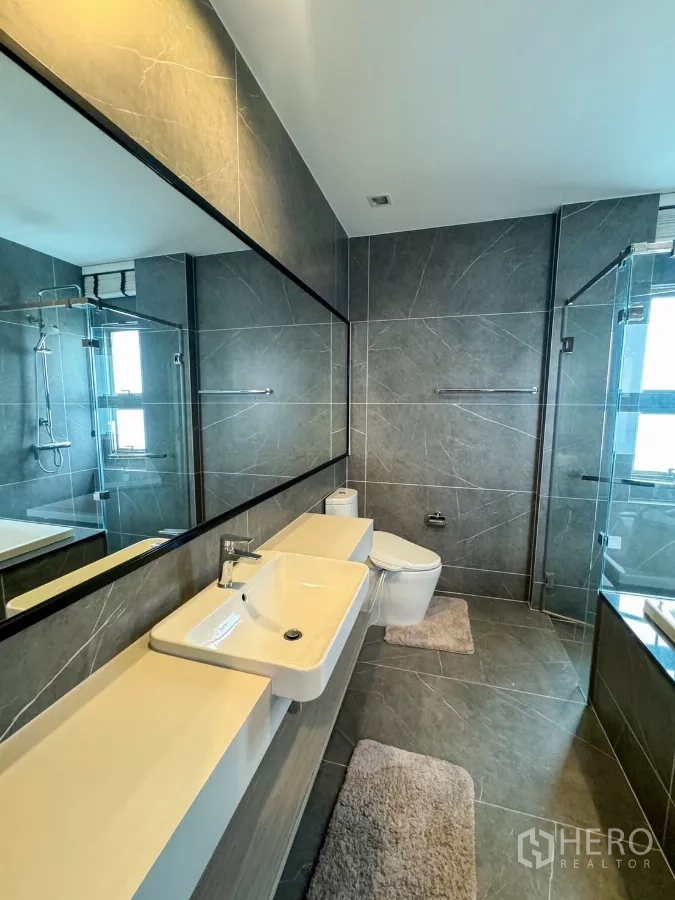 Detached House for rent or sale in Bueng Kum, Bangkok - Stylish bathroom with gray tiles, glass shower enclosure and long wall mirror.