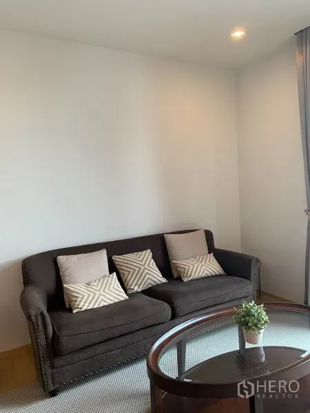 คอนโด ให้เช่าในPhaya Thai, Bangkok - ห้องนั่งเล่นอบอุ่น โซฟาสีเข้ม หมอนลาย และโต๊ะกระจกในคอนโดสว่าง