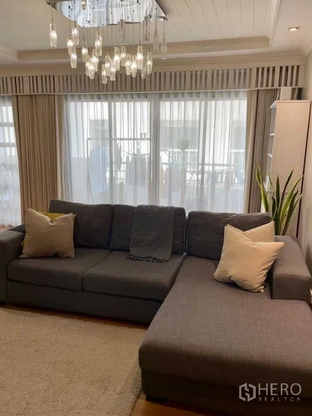 Condominium for sale in Watthana, Bangkok - Cozy living room with L-shaped gray sofa, chandelier and floor-to-ceiling curtains.