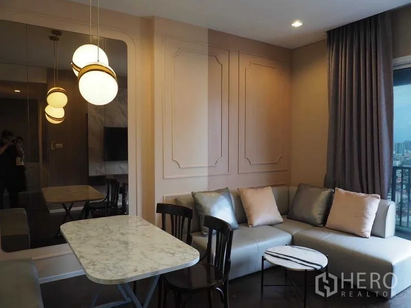 Condominium for rent in Chatuchak, Bangkok - L-shaped sofa and marble dining table in a bright living room with large window and balcony city view.