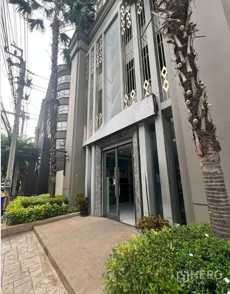 零售空间 出租在Khlong Toei, Bangkok - 零售空间入口，通高玻璃门、绿化花坛、人行道与棕榈树外立面，位于空堤区。