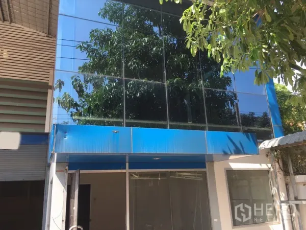 Warehouse for rent in Suan Luang, Bangkok - Modern glass-front entrance with blue canopy at the warehouse facade shaded by trees.