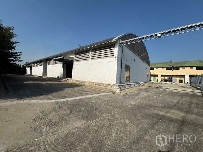 Warehouse for rent in Photharam, Ratchaburi - Exterior of white warehouse with curved metal roof and wide concrete yard for trucks.