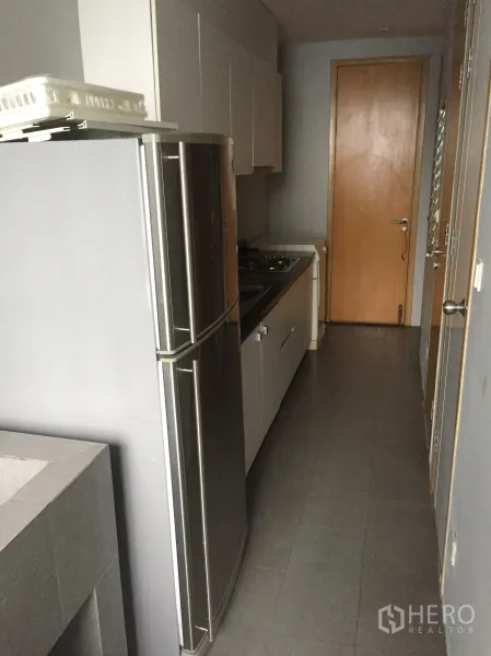 Condominium for rent or sale in Watthana, Bangkok - Compact galley kitchen with tall refrigerator, gas cooktop and a wooden door at the end.