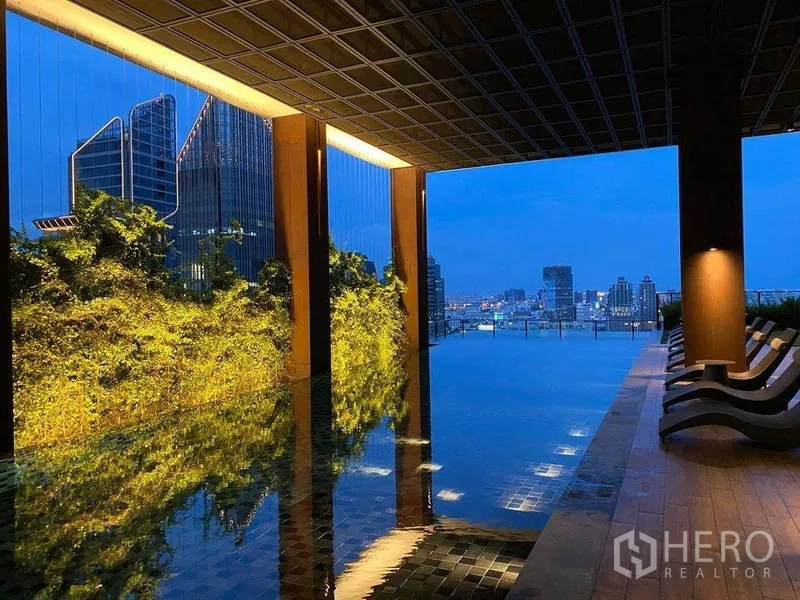 Condominium for rent or sale in Watthana, Bangkok - Rooftop infinity pool with city skyline and vertical garden wall, plus lounge chairs along a covered deck at dusk.