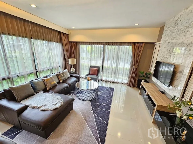 Detached House for rent in Prawet, Bangkok - Living room with L-shaped leather sofa, full-height windows, and sheer curtains.