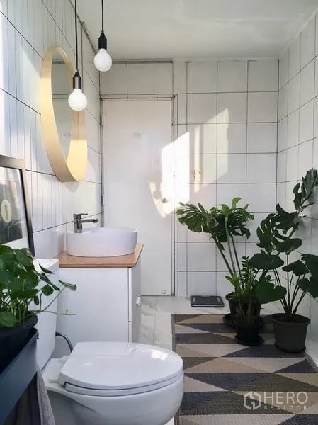Apartment for rent in Khlong Toei, Bangkok - Modern white-tiled bathroom with round mirror, vanity, toilet, pendant lights, and indoor plants.