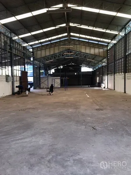 Warehouse for rent in Samut Prakan - Spacious clear-span warehouse floor with concrete slab under a metal truss roof allowing natural light.