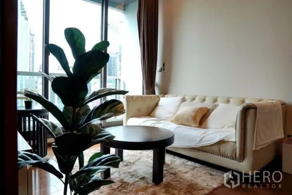 คอนโด ให้เช่าในKhlong Toei, Bangkok - ห้องนั่งเล่นสว่าง โซฟาหนังสีครีม โต๊ะกลม กระถางต้นไม้ และหน้าต่างบานใหญ่เห็นวิวเมือง