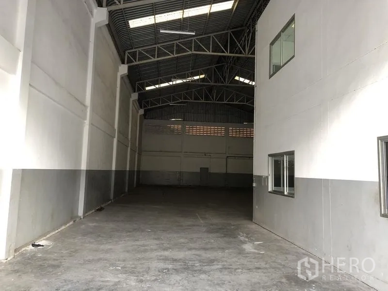 Factory for rent in Pathum Thani - Long warehouse aisle beside office block with high walls and concrete floor.