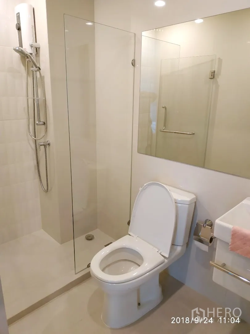 Condominium for rent in Huai Khwang, Bangkok - Modern bathroom featuring glass-enclosed shower, toilet and large mirror under recessed lighting.