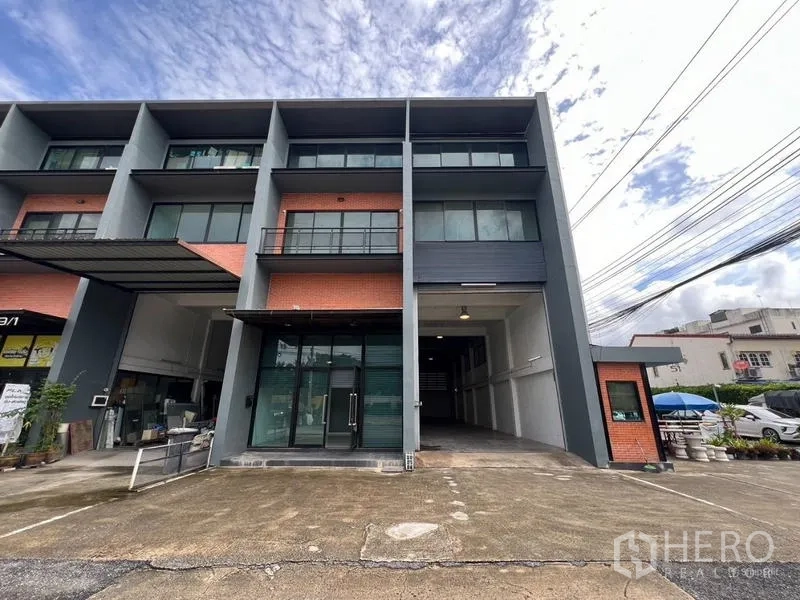 Warehouse for rent in Bang Phli, Samut Prakan - Modern three‑storey warehouse facade with brick accents, glass-fronted office, loading bay and parking under a cloudy sky.