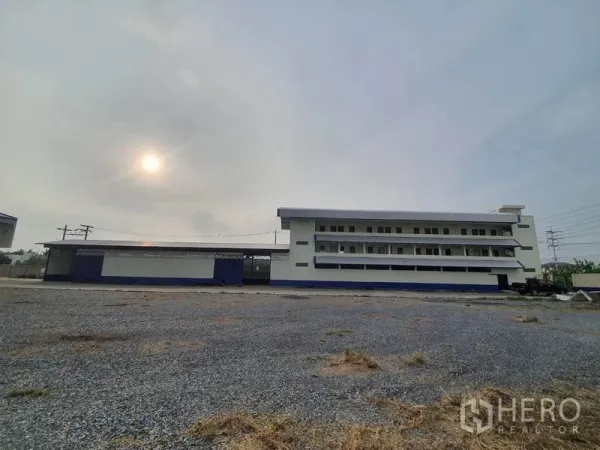 工厂 出租在Bang Bua Thong, Nonthaburi - 宽阔碎石场地与多层厂办楼外观，天空多云。