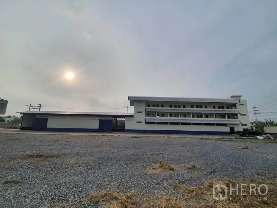 工厂 出租在Bang Bua Thong, Nonthaburi - 宽阔碎石场地与多层厂办楼外观，天空多云。