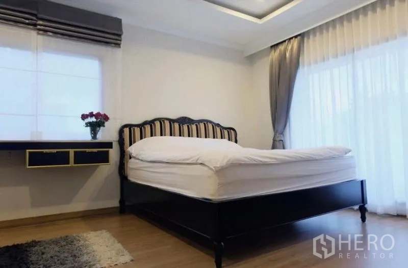 Detached House for rent in Suan Luang, Bangkok - Spacious bedroom with wood floors, large windows, sheer curtains, and a dark wood bedframe.