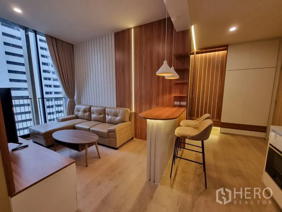 Condominium for rent in Watthana, Bangkok - Modern living area with L-shaped sofa, bar counter and stools, pendant lights, and floor-to-ceiling windows with blackout curtains and city view.