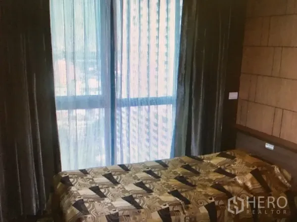 Condominium for rent in Ratchathewi, Bangkok - Second bedroom with bed, full-height window, blackout curtains and a city view through sheer drapes.