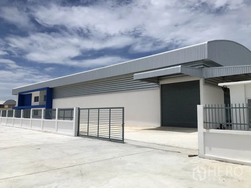 Warehouse for rent in Samut Prakan - Side view of warehouse showing wide concrete yard, sliding gate and a large roller shutter loading door.
