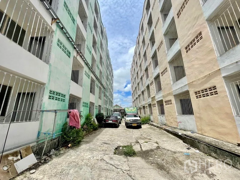 Apartment for sale in Mueang Samut Prakan, Samut Prakan - Two mid-rise apartment blocks with barred windows and a parking lane under blue sky.