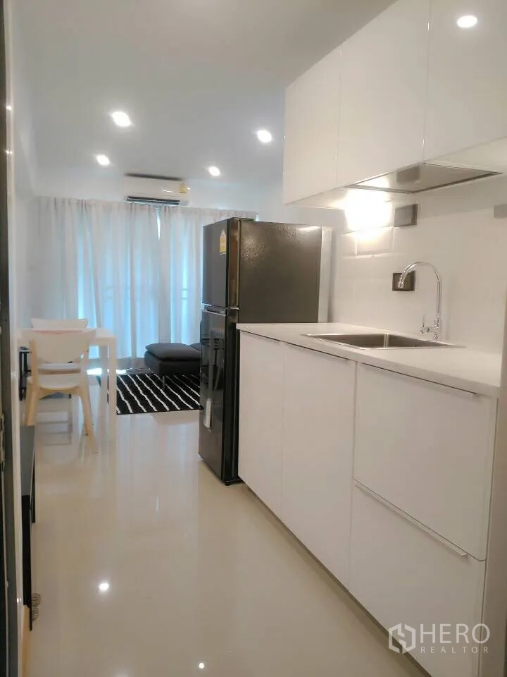 Condominium for rent in Watthana, Bangkok - Open-plan kitchenette with glossy white cabinets, stainless sink, large fridge, and dining set leading to the lounge.