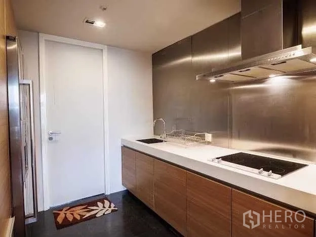 Condominium for sale in Yan Nawa, Bangkok - Modern closed kitchen with wood cabinetry, white countertop, dual induction hobs and stainless‑steel backsplash.