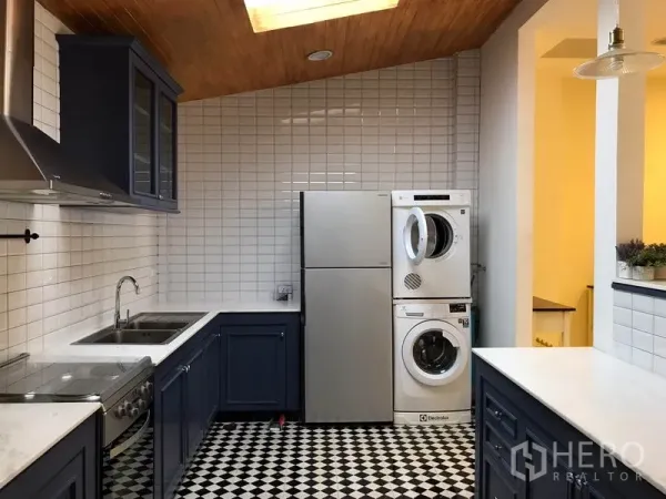 Townhouse for rent in Wang Thonglang, Bangkok - Kitchen corner showing fridge and stacked washer–dryer beneath a skylight and white tile walls.