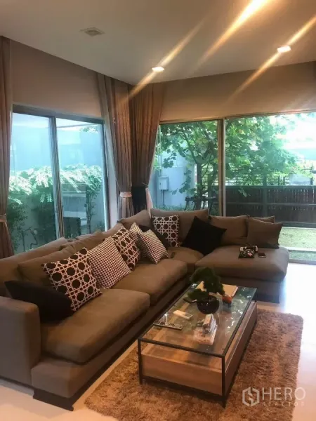 Detached House for rent in Bang Kapi, Bangkok - Bright living room with large sectional sofa, floor-to-ceiling windows and garden view.