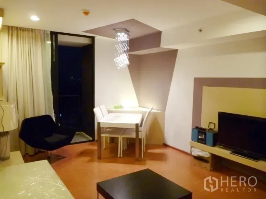 Condominium for rent in Watthana, Bangkok - Open-plan living and dining area with balcony access, TV feature wall and modern chandelier.