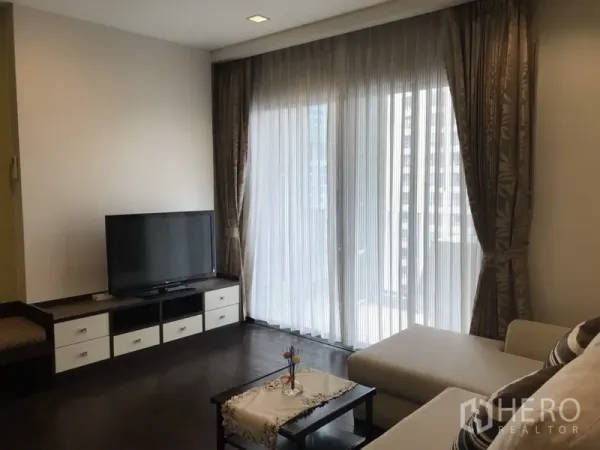 Condominium for rent in Ratchathewi, Bangkok - Bright living room with TV console, dark wood floors and floor-to-ceiling sheer curtains opening to a balcony city view.