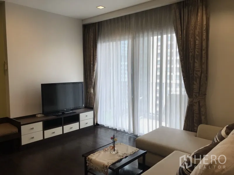 Condominium for rent in Ratchathewi, Bangkok - Bright living room with TV console, dark wood floors and floor-to-ceiling sheer curtains opening to a balcony city view.