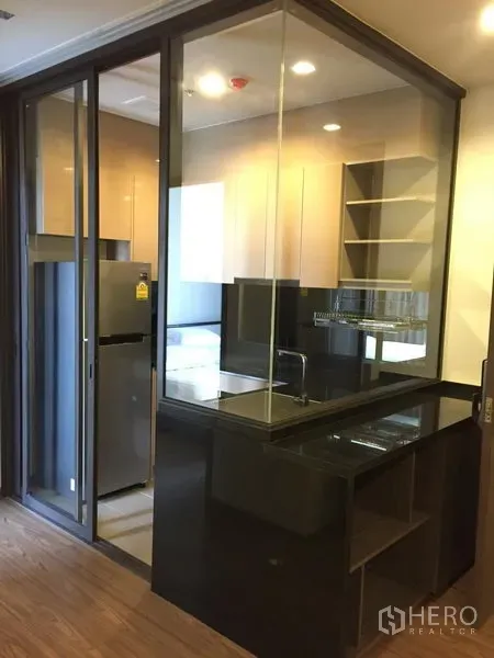 Condominium for rent in Watthana, Bangkok - Glass‑enclosed kitchenette with black countertops, sink, shelving and a stainless refrigerator.