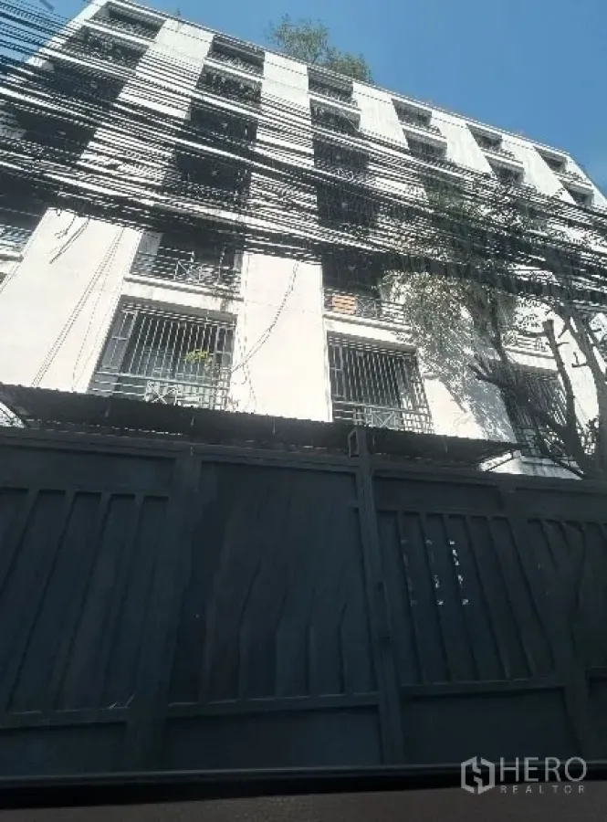 คลังสินค้า ให้เช่าในKhlong Toei, Bangkok - มุมมองด้านหน้าประตูโกดังคอนกรีตพร้อมเหล็กดัด ปกคลุมด้วยร่มเงาต้นไม้ และสายไฟหนาแน่น ใจกลางกรุงเทพฯ