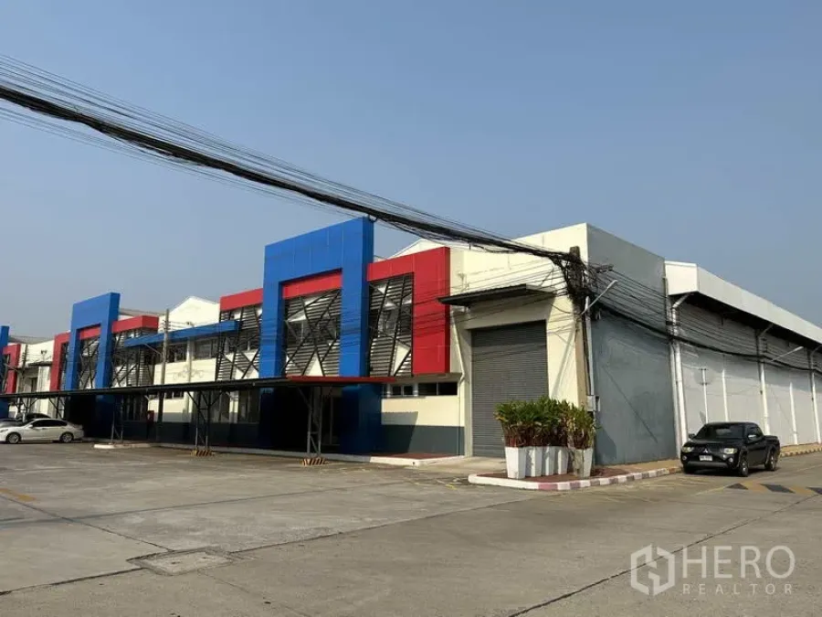 โรงงาน ให้เช่าในBang Phli, Samut Prakan - มุมมองด้านหน้าโรงงานสมัยใหม่ โทนสีน้ำเงิน–แดง มีที่จอดรถและประตูม้วนขนาดใหญ่