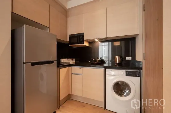 Condominium for rent in Khlong Toei, Bangkok - Modern L-shaped kitchen with light wood cabinets, black countertop, fridge, cooktop, microwave, and washing machine.