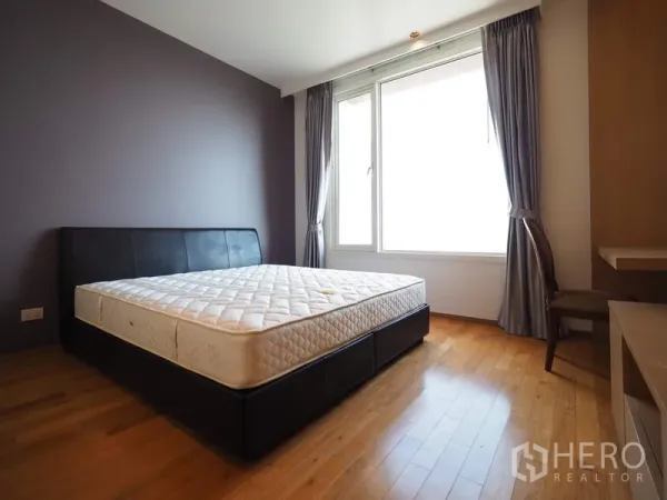 Condominium for rent in Sathon, Bangkok - Sunlit bedroom with large window, gray curtains, wood floors and an upholstered bed frame.