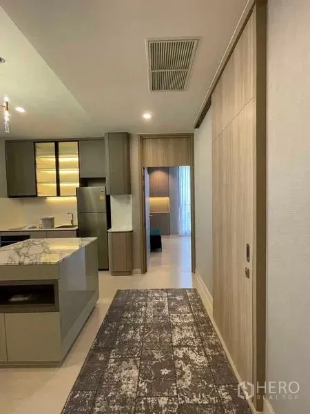 Condominium for rent in Pathum Wan, Bangkok - Hallway leading to an open kitchen with marble island, built-in cabinets and a stainless-steel fridge.
