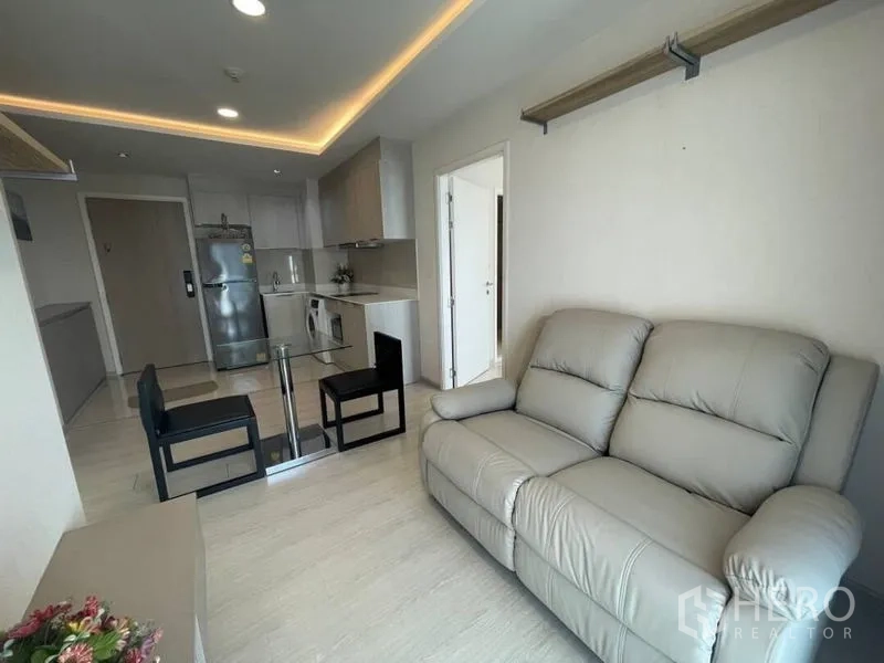 Condominium for rent in Khlong Toei, Bangkok - Bright living room with grey leather sofa, glass dining table, and open kitchen with fridge and washer.