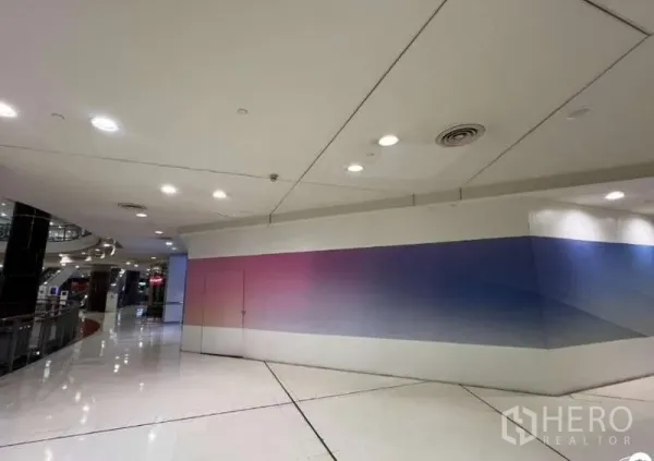 พื้นที่ขายปลีก ให้เช่า - พื้นที่หน้าร้านมุมในโถงศูนย์การค้า พื้นสีขาวเงา เพดานฝ้าพร้อมไฟดาวน์ไลท์ และผนังสำหรับติดแบรนด์ยาว