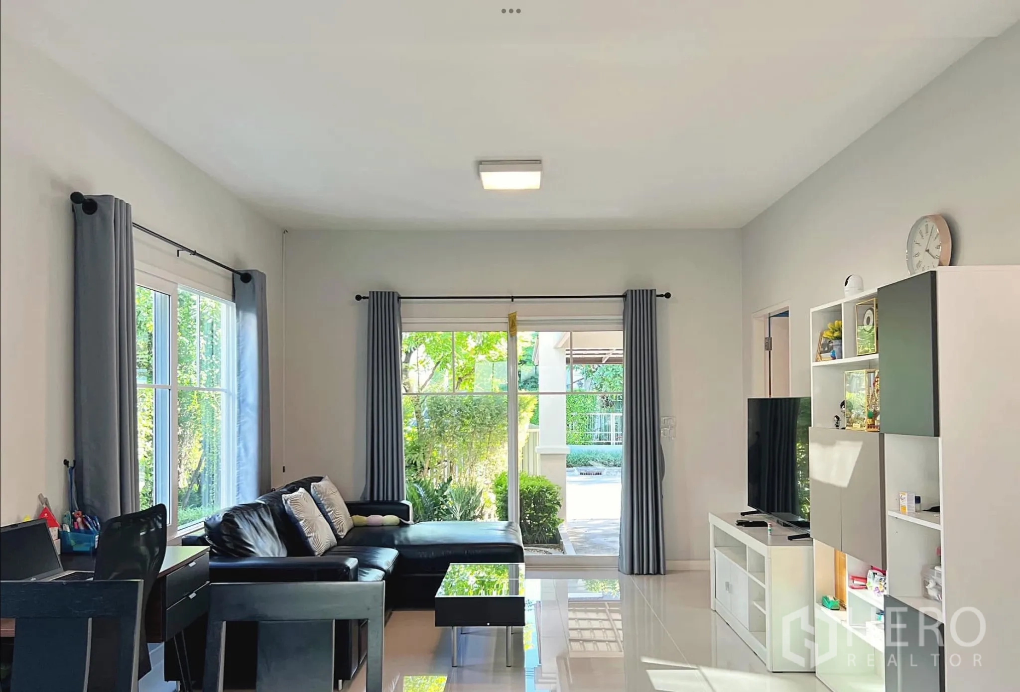 Townhouse for rent or sale in Sai Mai, Bangkok - Bright living room with large windows, grey curtains, TV unit and garden views.