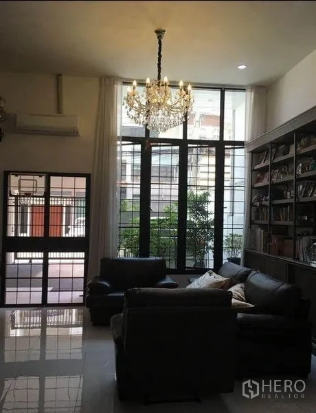 Townhouse for rent in Bang Rak, Bangkok - High-ceiling living room with chandelier, tall windows, leather sofas, and a large bookcase.