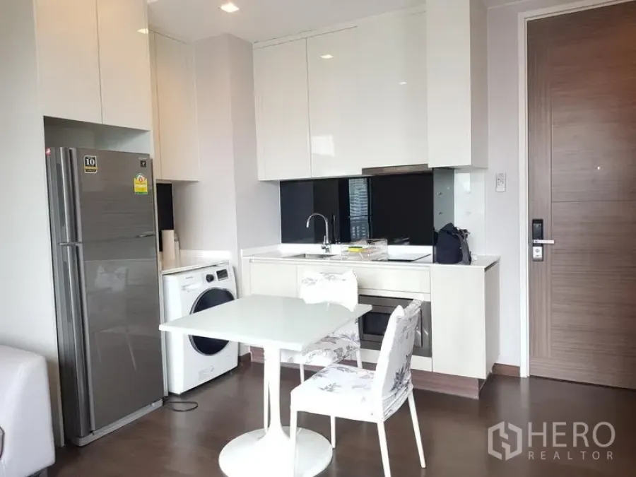 公寓 出租在Ratchathewi, Bangkok - 现代一体化小厨房，白色亮面柜体、嵌入式烤箱、冰箱与洗衣机，旁有餐桌。